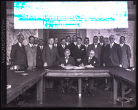 Two men sign documents with a group of men behind them, Los Angeles, 1930s