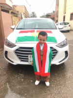 Voting in Khanaqin, A young boy wearing Kurdish clothing, flag of Kurdistan around his neck, and standing in front of a white car with a flag of Kurdistan on the hood