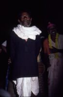 Mannan man looks into the camera with the dancer Perumal behind him at a festival of the Mannan Ādivāsī people, Mannakudi (Tamil Nadu, India), 1984