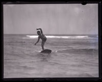 Surfer and Olympic swimmer Duke Kahanamoku standing on a surfboard on one leg, Los Angeles, 1920s