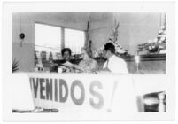 Guadalupe Kirarte Domínguez sentada en una mesa con un letrero que dice "Bienvenidos!"