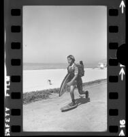 David Dollase, with surfboard under arm skateboarding along the beach near Santa Monica Canyon, Calif., 1975