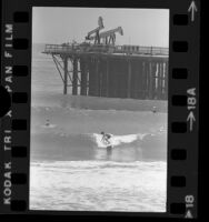 Surfers riding waves with offshore oil wells in the background near Santa Barbara, Calif., 1976