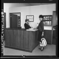 Woman at ticket counter modeling travel clothing, brown and black mohair skirt and jacket, Los Angeles, Calif., 1962
