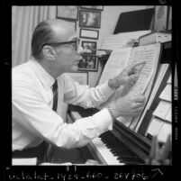 Conductor John Green seated at piano examining sheet music, 1970