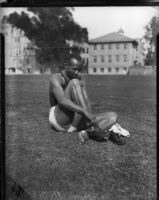 Jesse Owens puts on running shoes while sitting on grass, Los Angeles, 1930s