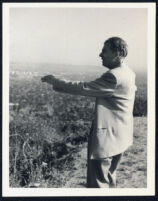 Aldous Huxley, arm outstretched, standing in hills above Hollywood [descriptive]