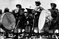 Photo of panpipe ensemble, with two men playing caja (large drum), and multiple panpipe players
