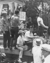 Men play barrel drums atop a parade float; another man displays a framed photograph of Nai Tun playing the phia