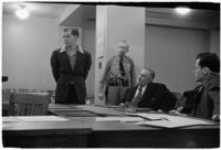 Man testifying (?) during unidentified hearing in courtroom with model cars on table