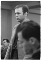 Man testifying (?) during unidentified hearing in courtroom with model cars on table