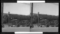 Two cars parked at curb in front of a walkway and (adobe?) building (unidentified), California, circa 1915-1925