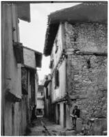Man sitting in front of a stone building next to a narrow alley, Nérac, France, 1929