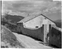 Mountain road running behind a small house and gate in a concrete wall, Europe, 1929
