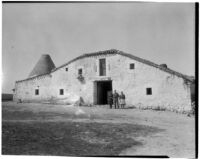 Man and two women standing in front of a large stone and stucco barn in a field, Spain, 1929