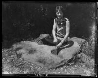 Grinding stone with 3 hollows, on ground among foliage, with Clarice Barclay seated demonstrating grinding, near Saddle Peak, Los Angeles, 1929