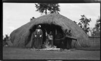 Tribal chief regally seated in the doorway of a traditional round dwelling with guard and drummers, Uganda, 1933
