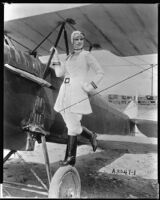 Peggy Hamilton standing on the wheel of an airplane and modeling an aviation costume created especially for her by Greer, 1928