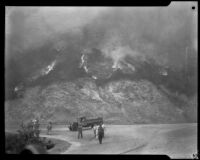 Fire fighters walk down a road as fire consumes brush on the hillside behind them in Griffith Park, Los Angeles, 1929