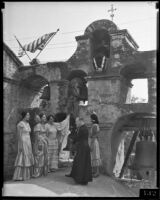 Priest and people in Spanish dress beside bells of Misison San Gabirel, 1920-1939
