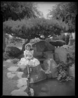 Helena Burnett in dress and bonnet sitting next to a pond, 1947-1950