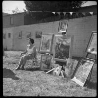 Artist seated next to a display of her paintings at a Santa Monica Art Association exhibition, Santa Monica, 1962