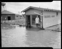 Barber standing in front of his shop on a rain-flooded street, Compton, [1927?]