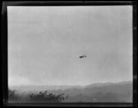 Airplane in sky above tree tops and mountains, circa 1920
