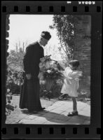 Father Arthur John Hutchinson gives a small girl a basket of flowers at mission San Juan Capistrano, 1936
