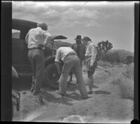 University student and 2 men watch a man repair a punctured tire on a tow car, Morongo Valley, 1933