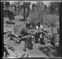 Neil Wells and H. H. West, Jr. sit in the middle of a woodpile outside a cabin, Big Bear, 1932