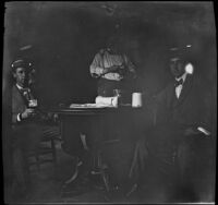 Two men sit at a table holding drinks while another man stands behind them, Burlington, 1900