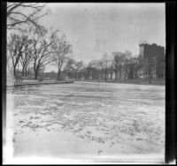 Frozen pond with trees, buildings, and benches in the background, Boston, 1914