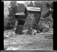 H. H. West, Jr. and Mertie West sit on snow-covered rocks outside a large rustic-style house, Redlands vicinity, about 1930