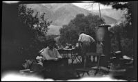 William Shaw sits at a campsite while three others (probably Mertie West and Agnes and Forrest Whitaker) stand in the background, Inyo County vicinity, about 1930