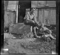 Roy Bagley sitting outside his cabin and putting on boots, Westlake Village, about 1915