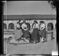 Bessie Velzy sits on a fountain with three other women across the street from Mission San Fernando Rey de España, Mission Hills, Los Angeles, 1909