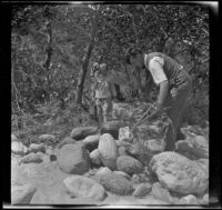 Glen Velzy cooks while Frances West carries a pail and Elizabeth West watches, San Gabriel Canyon, about 1915