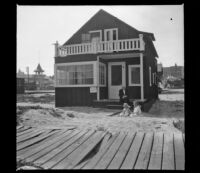 H. H. Cooper sits on the porch of the West's beach cottage while Elizabeth and Frances West sit in the sand in front, Venice, about 1910