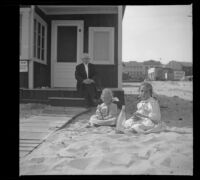 H. H. Cooper sits on the porch of the West's beach cottage while Elizabeth and Frances West sit in the sand in front, Venice, about 1910