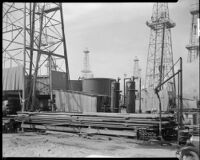 Stack of casing in front of an oil derrick, probably at the Venice oil field, Los Angeles, 1930
