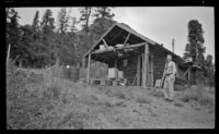 View of the West's bus driver posing next to Pioneer cabin at Igloo Creek, Denali National Park and Preserve, 1946