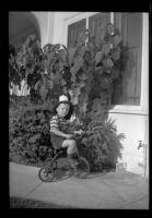 Tommy Newquist wears a fireman's helmet while riding a tricycle in front of the West residence, Los Angeles, 1948