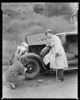 Fred Keating, actor, seated on the running board of a car in the Hollywood Hills as a woman changes the tire, Los Angeles, circa 1934