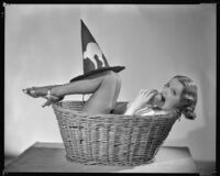 Inez Courtney, actress, in a basket biting an apple with witch's hat on her legs, circa 1934-1939