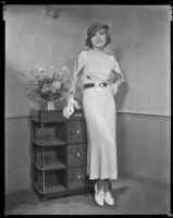 Woman wearing a hat, dress, and gloves and leaning on a dresser with a hand on her hip, circa 1926-1939