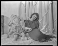 Barbara Read, actress, with her eyes closed and holding the hand of a large doll, circa 1934-1936