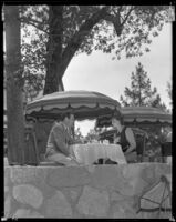Johnny Mack Brown, actor, holding out a basket to a woman as they sit at an outdoor table, Lake Arrowhead, circa 1929-1934