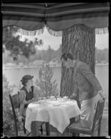 Johnny Mack Brown, actor, pulling out a chair from a table where a woman sits, Lake Arrowhead, circa 1929-1934