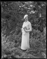 Woman standing among trees while holding a purse and wearing a hat, circa 1932-1939